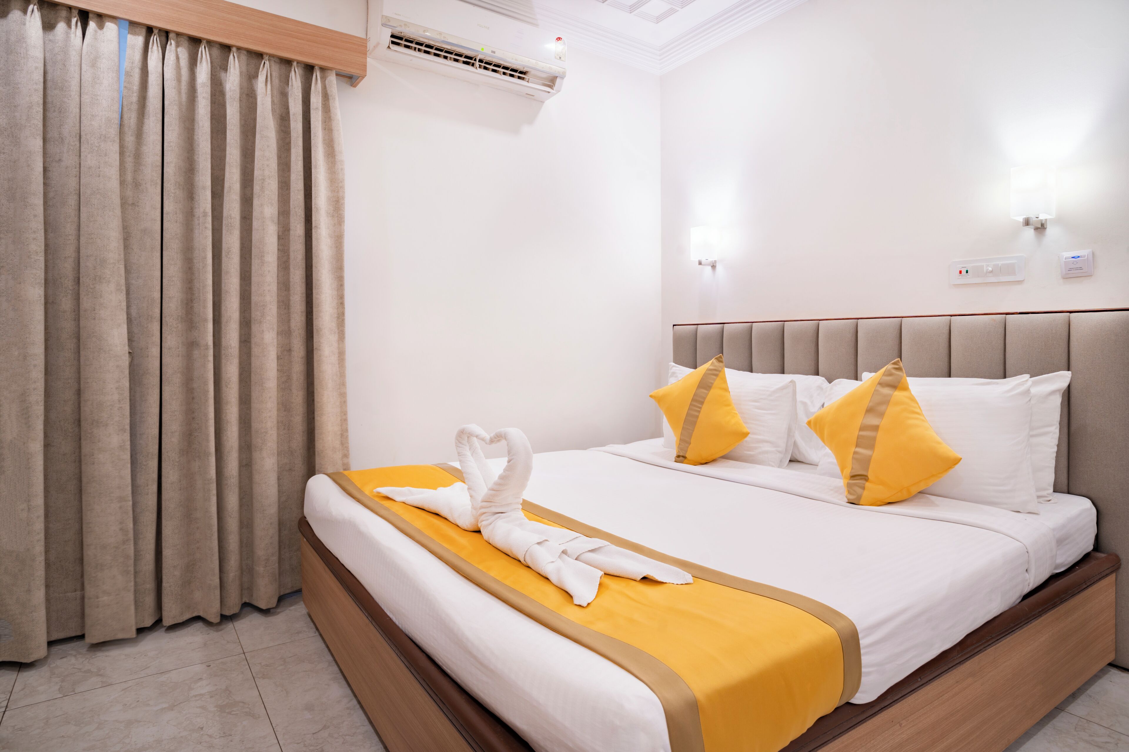 Una habitación de hotel con cama, cortinas, un equipo de aire acondicionado y una lámpara fijada a la pared.