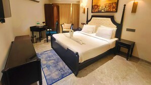 Habitación de hotel con cama, escritorio, silla, lámpara de pie, ventilador de techo y un cuadro en la pared.