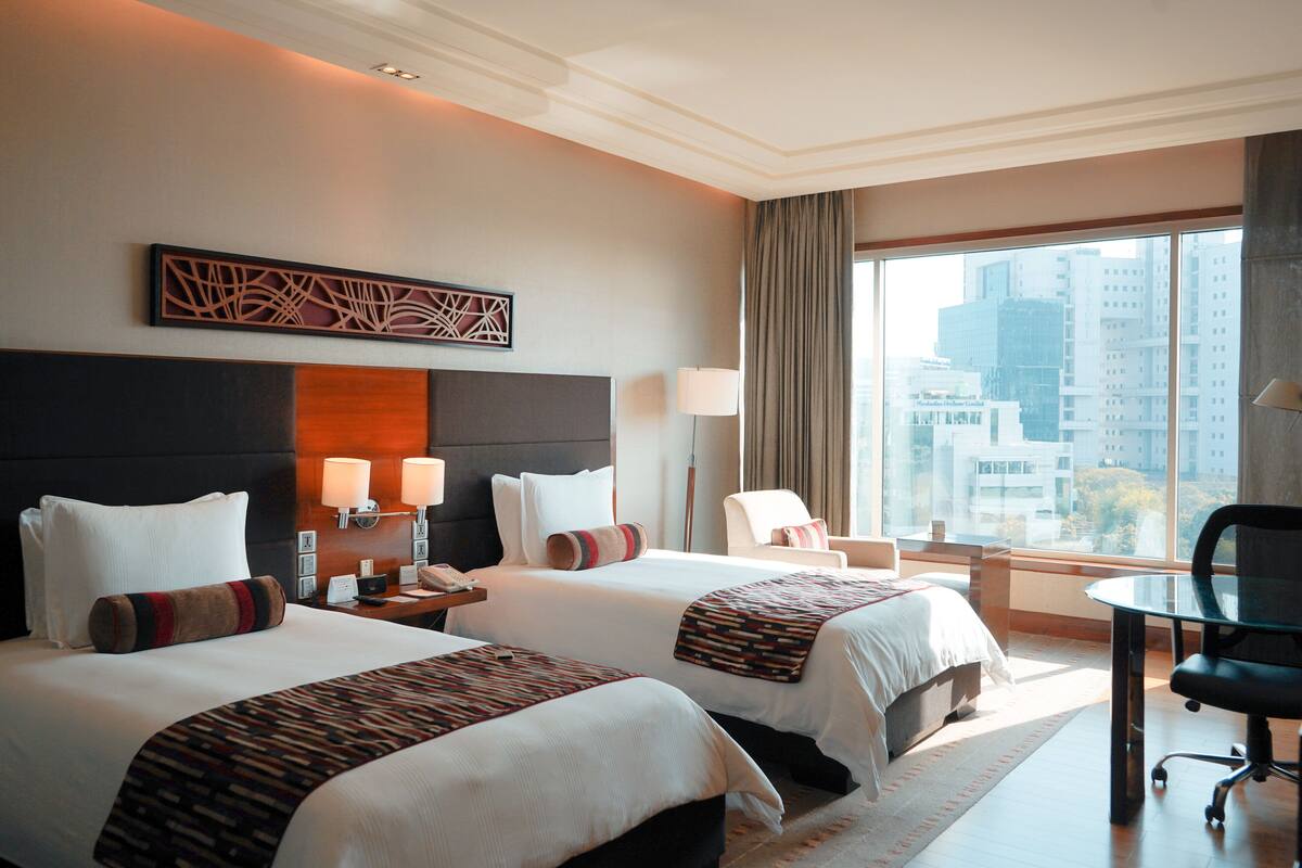 A hotel room with two beds, a desk, and a large window offering a city view.