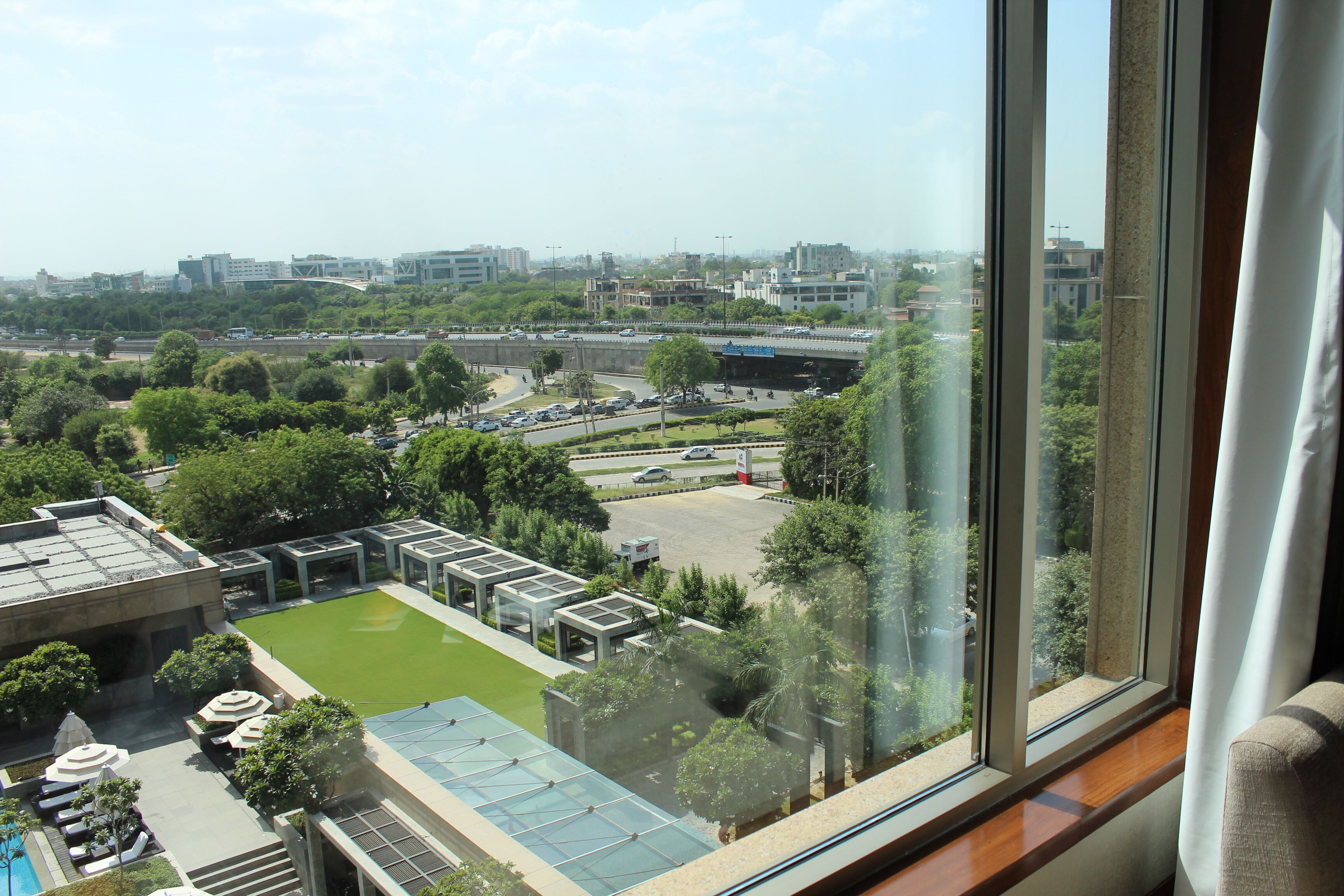 Vue depuis une fenêtre d’hôtel, donnant sur une cour aménagée avec une piscine, un parking et, au loin, le paysage urbain.