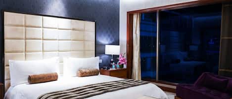 A hotel room with a large bed, a nightstand, a bench, a purple armchair, and a view of the city through a large window.