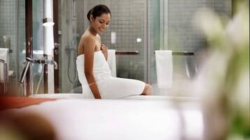 A woman sitting in a bathtub in a modern bathroom with glass walls and a visible shower area.