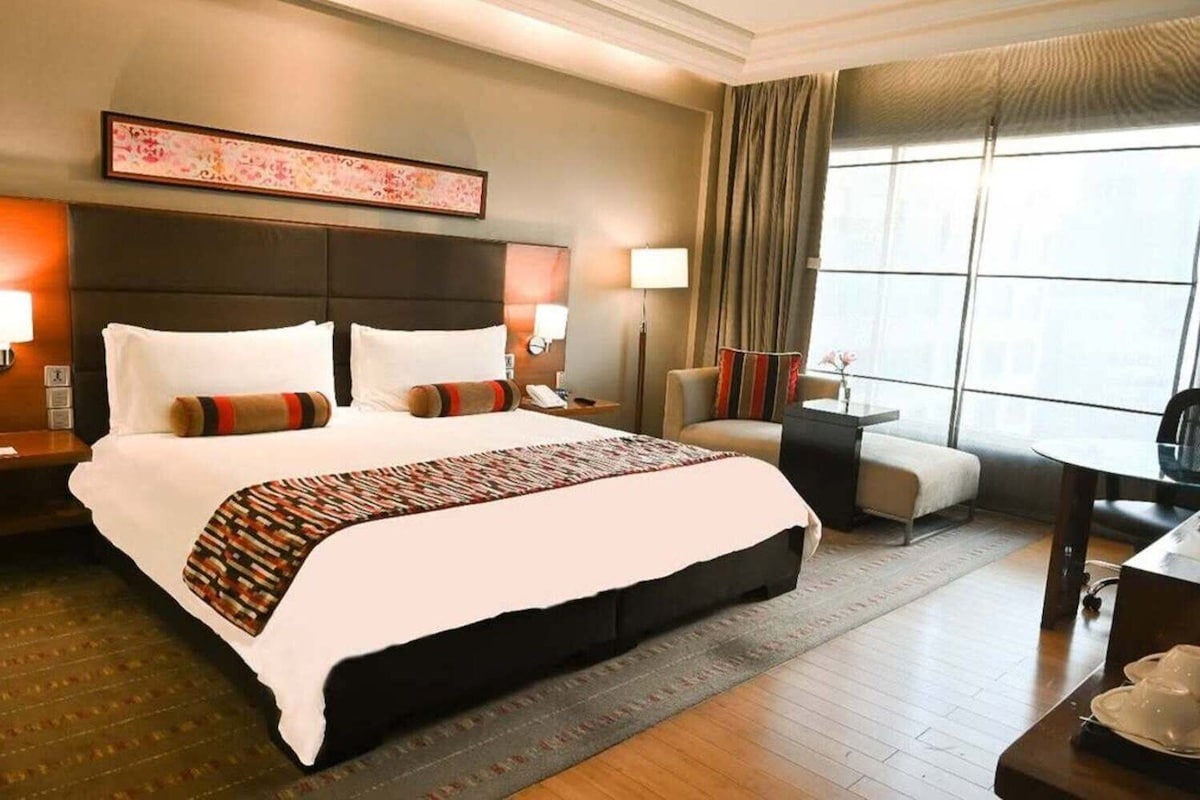 A hotel room with a large bed, a desk, a chair, a nightstand, and a window with curtains.
