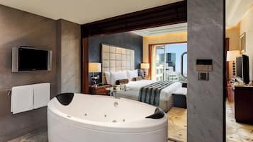 A hotel room with a large bathtub, a bed with a headboard, a television mounted on the wall, and a view of the cityscape through the window.