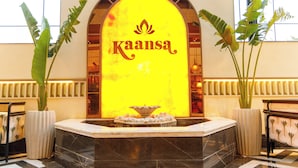 Un área de recepción con un cartel iluminado grande que dice "Kaansa", una fuente de mármol y plantas en macetas.
