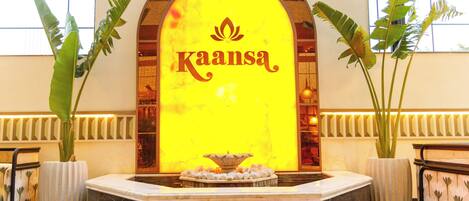 A reception area with a large illuminated sign reading "Kaansa," a marble fountain, and potted plants.