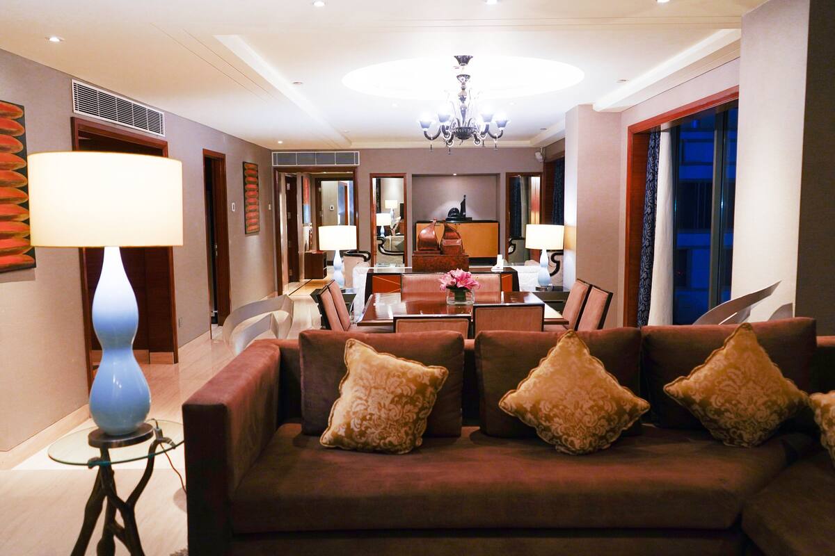 A modern hotel lobby with a sofa, lamps, a chandelier, and a dining area.