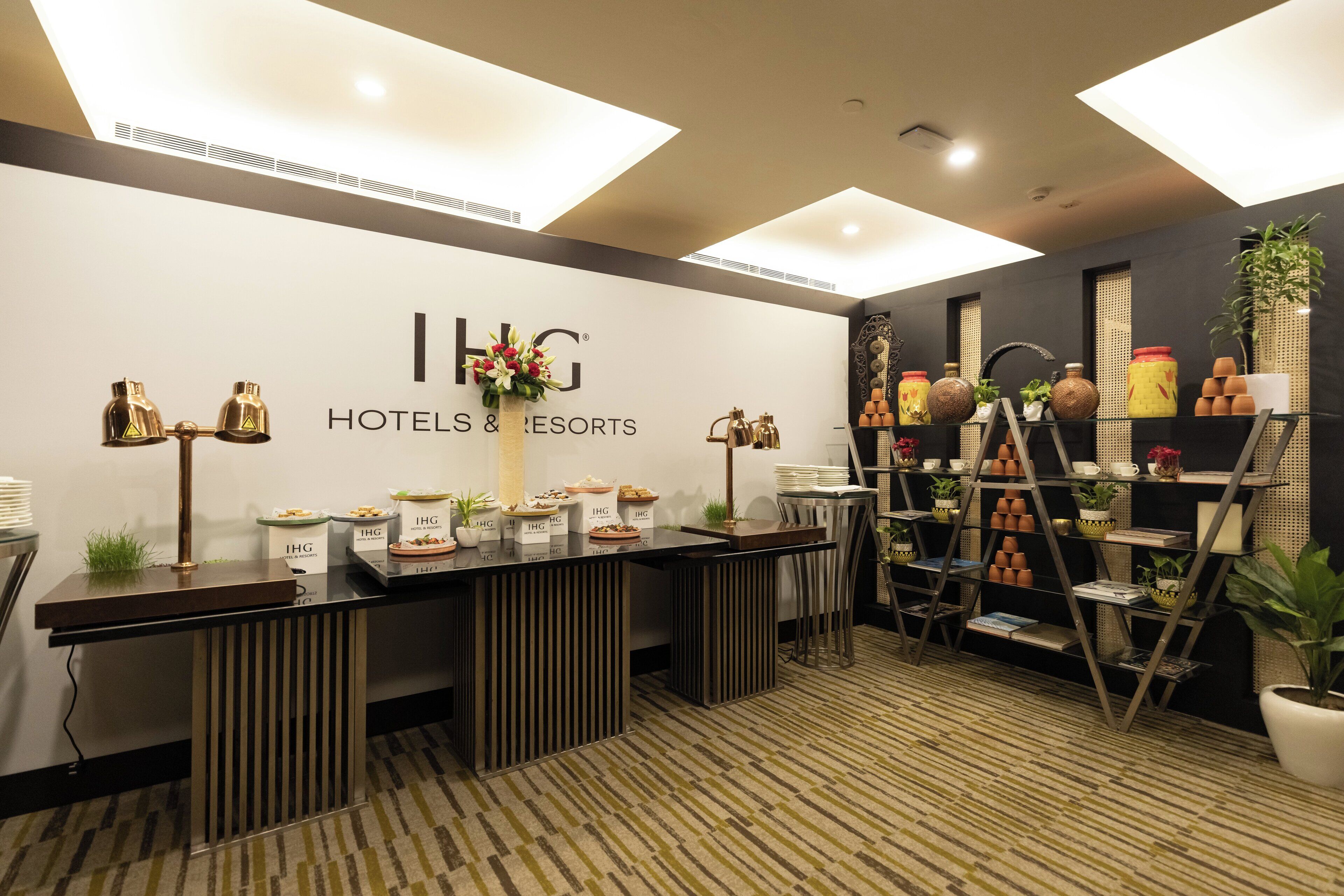 A reception area with a white wall displaying the logo and name of IHG Hotels & Resorts. There are buffet stations with labeled containers, a reception desk, and a display of decorative items and plants.
