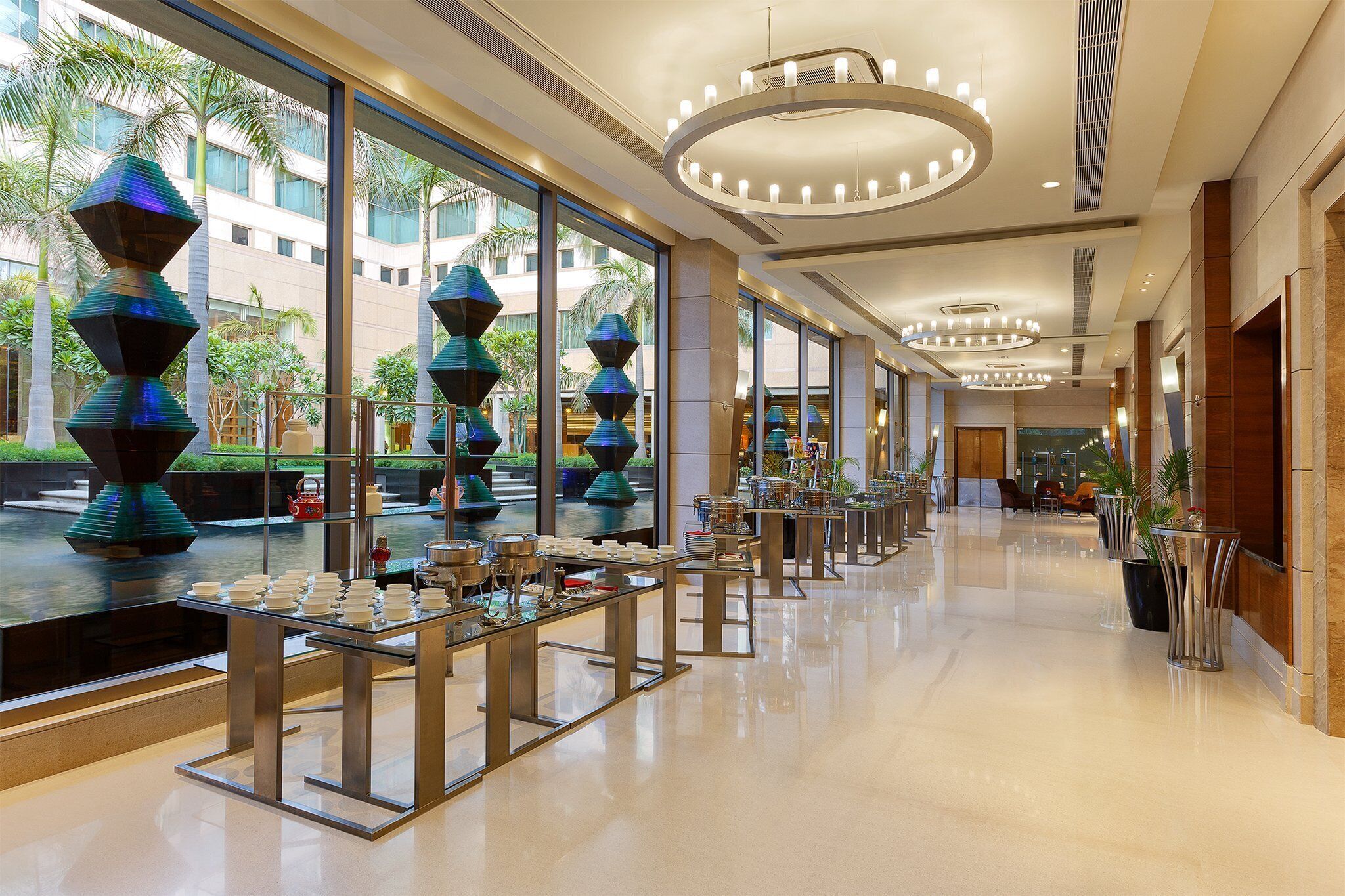 A hotel lobby with a long reception desk, modern chandeliers, and large windows offering views of palm trees and a building.