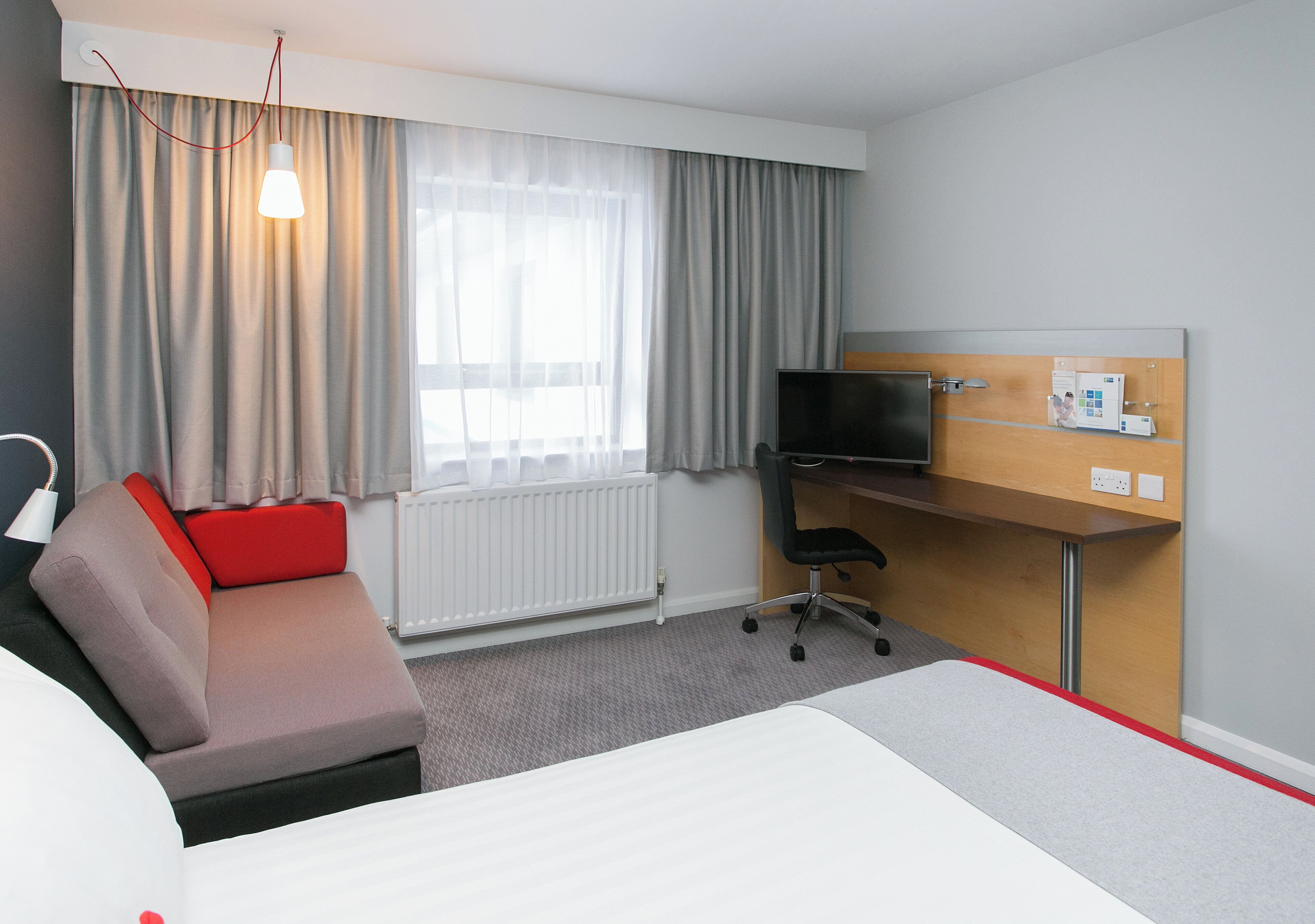 Habitación de hotel con una cama, un escritorio con computadora, una silla, un televisor y una ventana con cortinas.