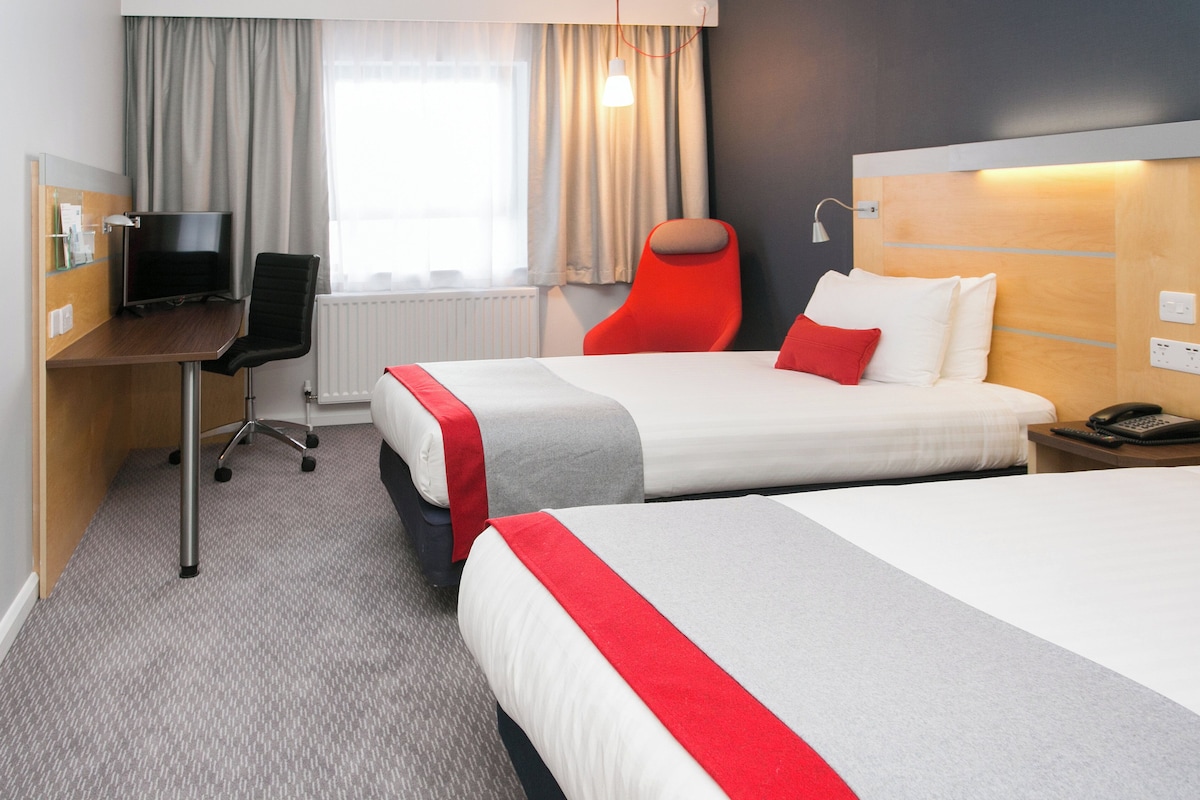 Ett hotellrum med två sängar, ett skrivbord med en stol, en tv och ett fönster med gardiner.