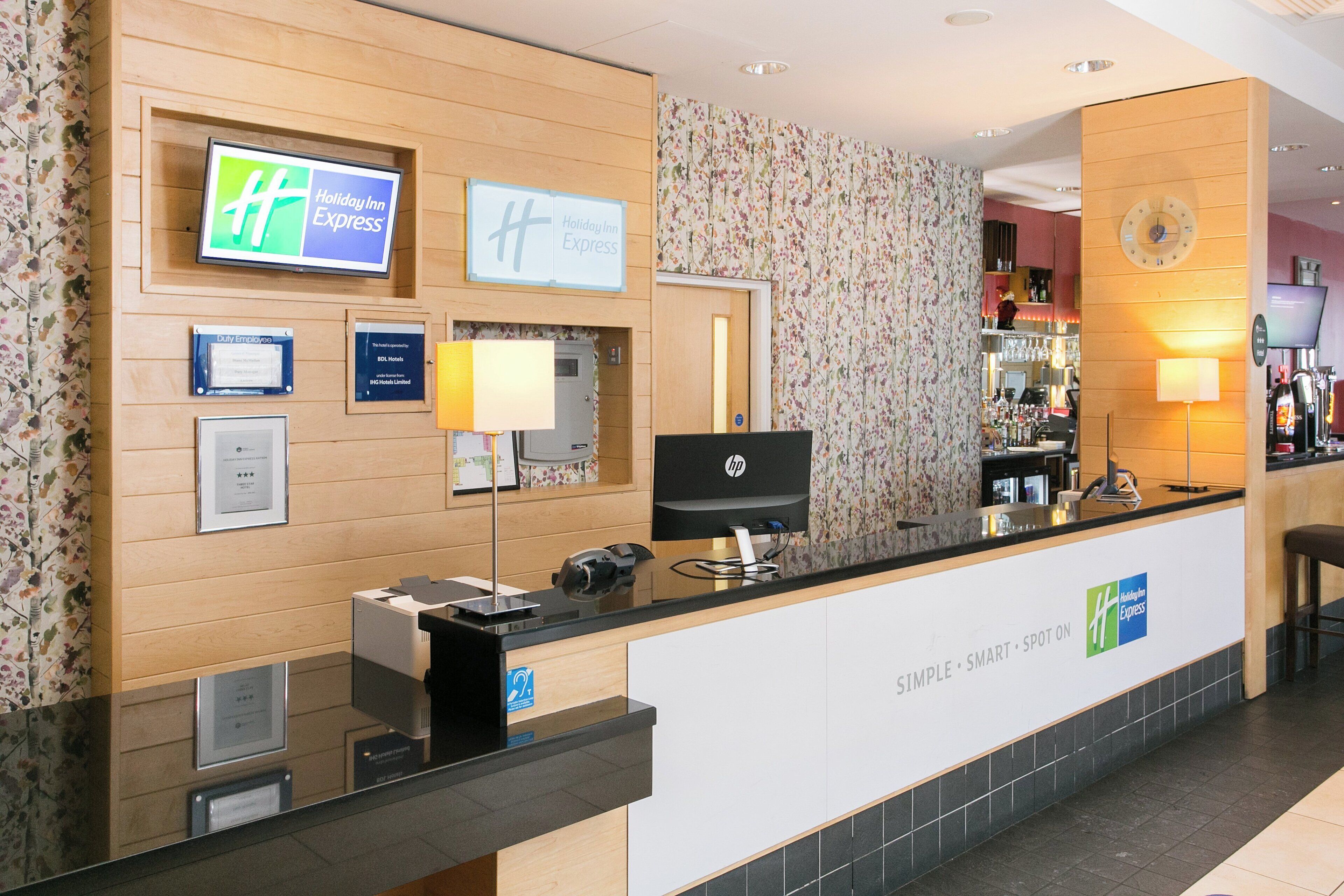 A hotel reception desk with a digital display, a computer, and a telephone. There are framed certificates and a clock on the wall.