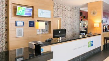 A hotel reception desk with a digital display, a computer, and a telephone. There are framed certificates and a clock on the wall.