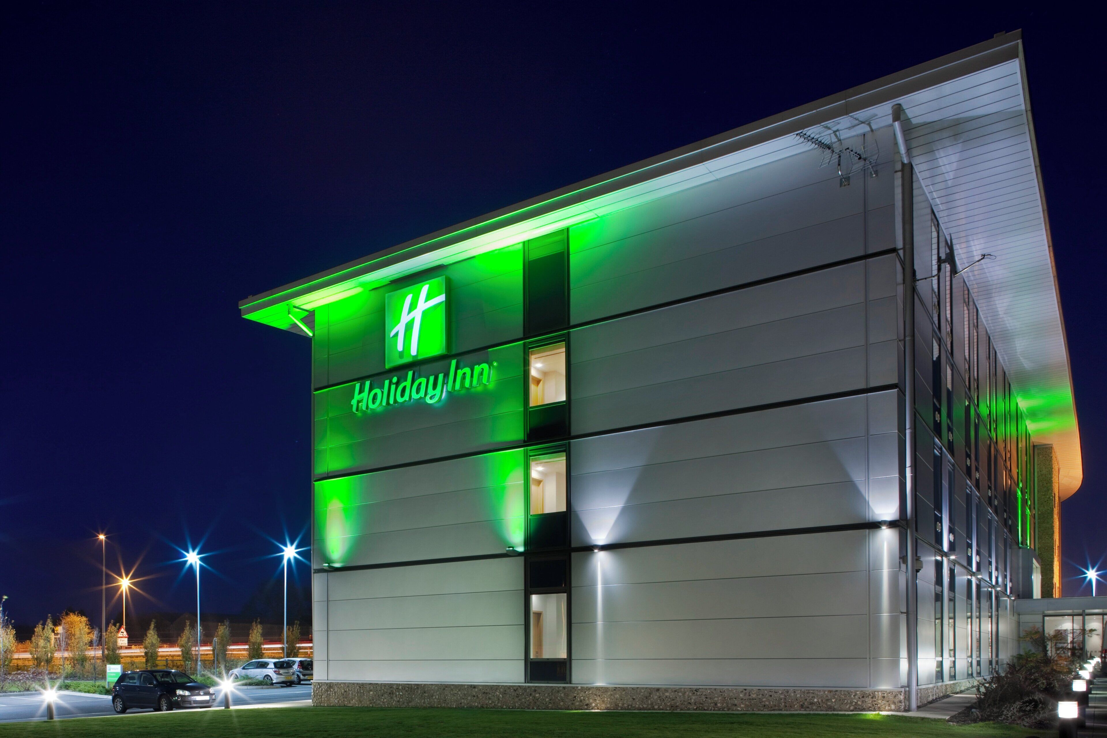 Un hotel Holiday Inn con fachada moderna, señalización iluminada y un estacionamiento con automóviles.