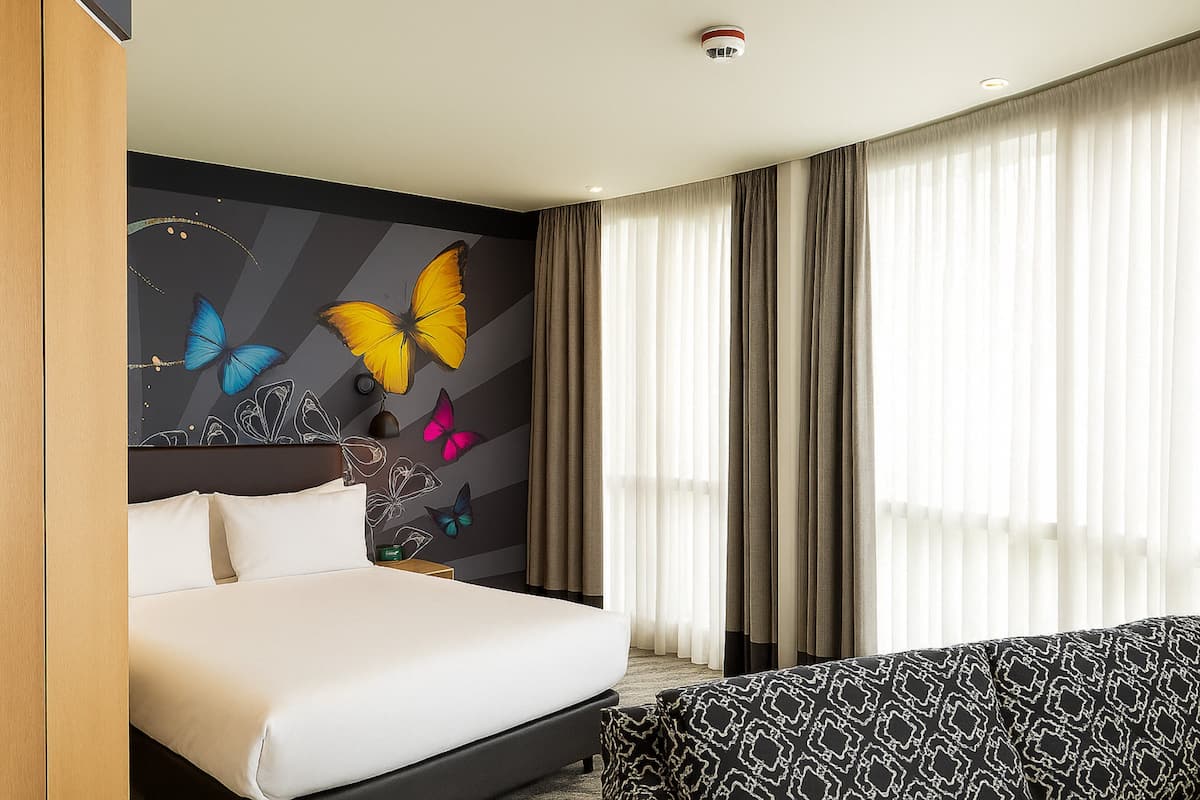 A hotel room with a large bed, a wall mural with butterflies, a window with sheer curtains, and a patterned sofa.