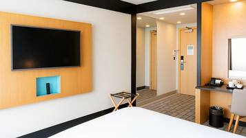 A hotel room with a bed, a TV mounted on the wall, a desk with a chair, a trash bin, and a mirror.
