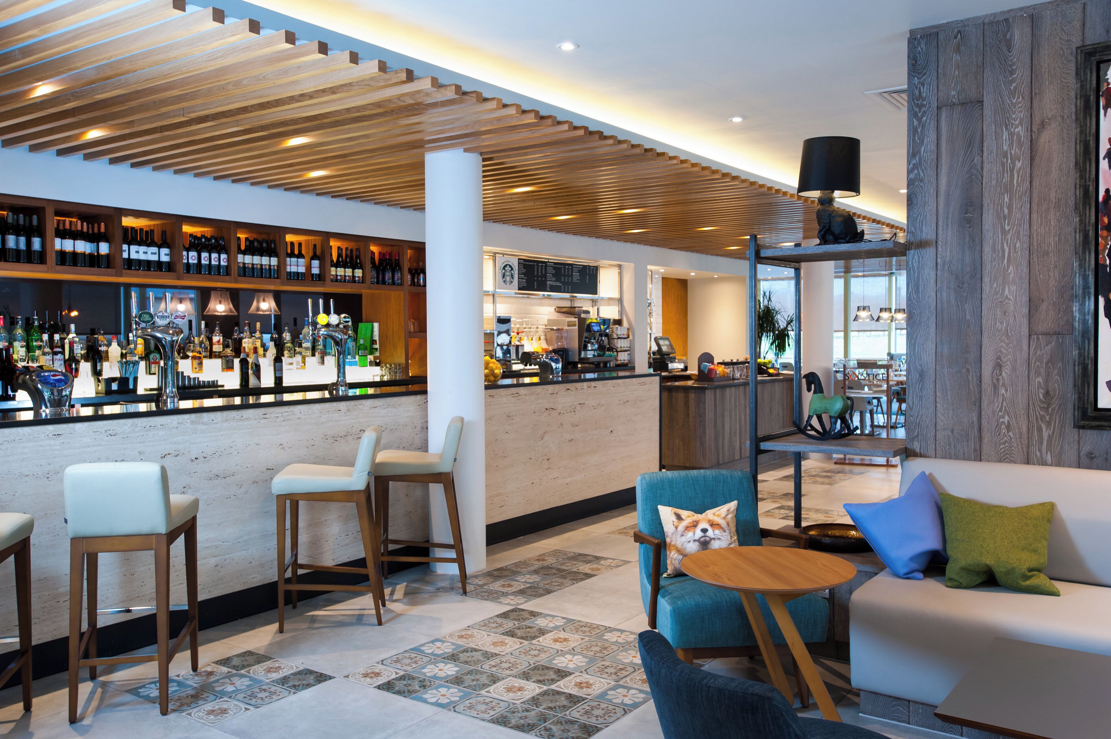 Een moderne barruimte met een houten plafond, zitplaatsen met kussens en een diverse collectie flessen op planken.