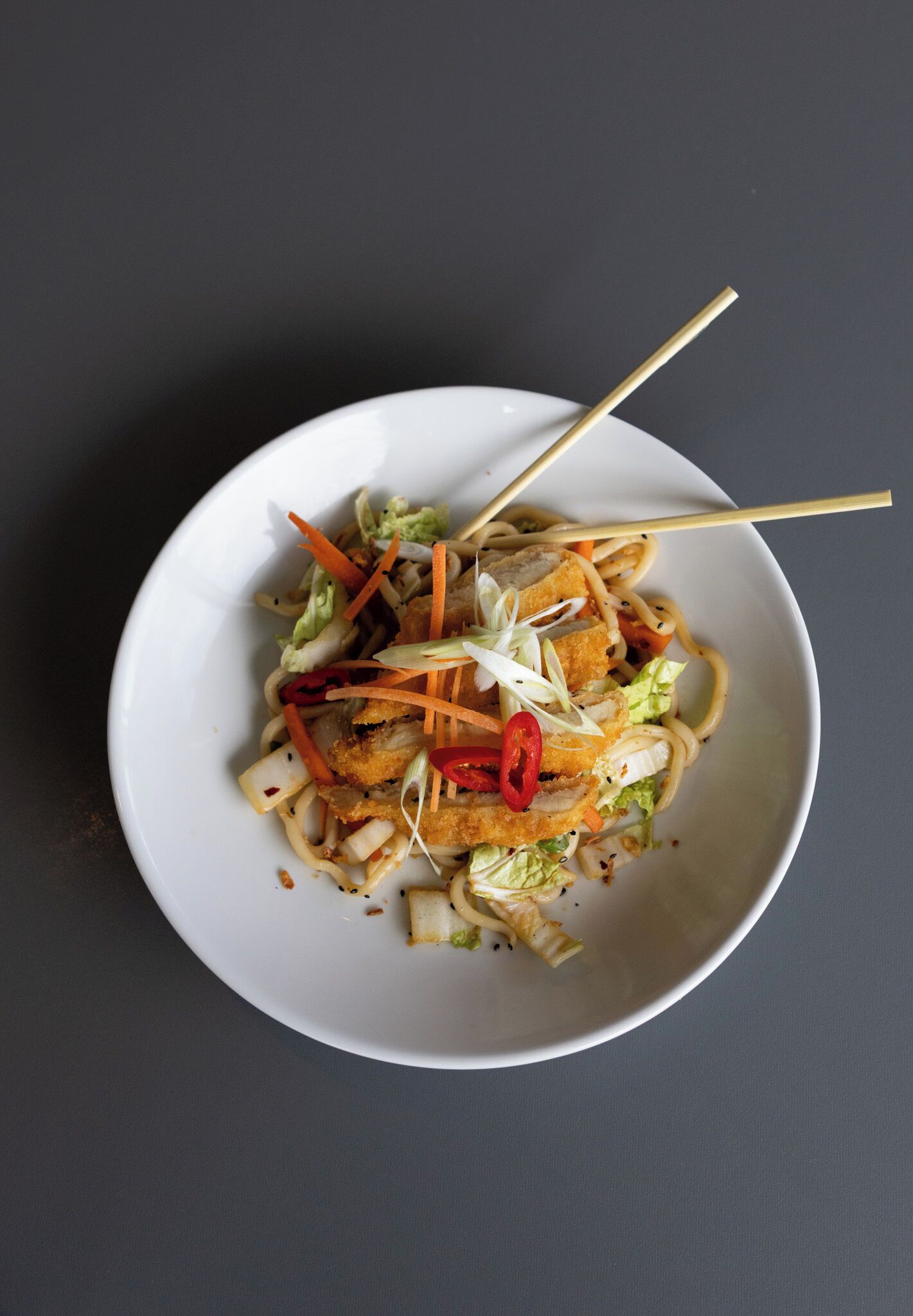 A bowl of noodles with vegetables and meat, served on a white plate with chopsticks.