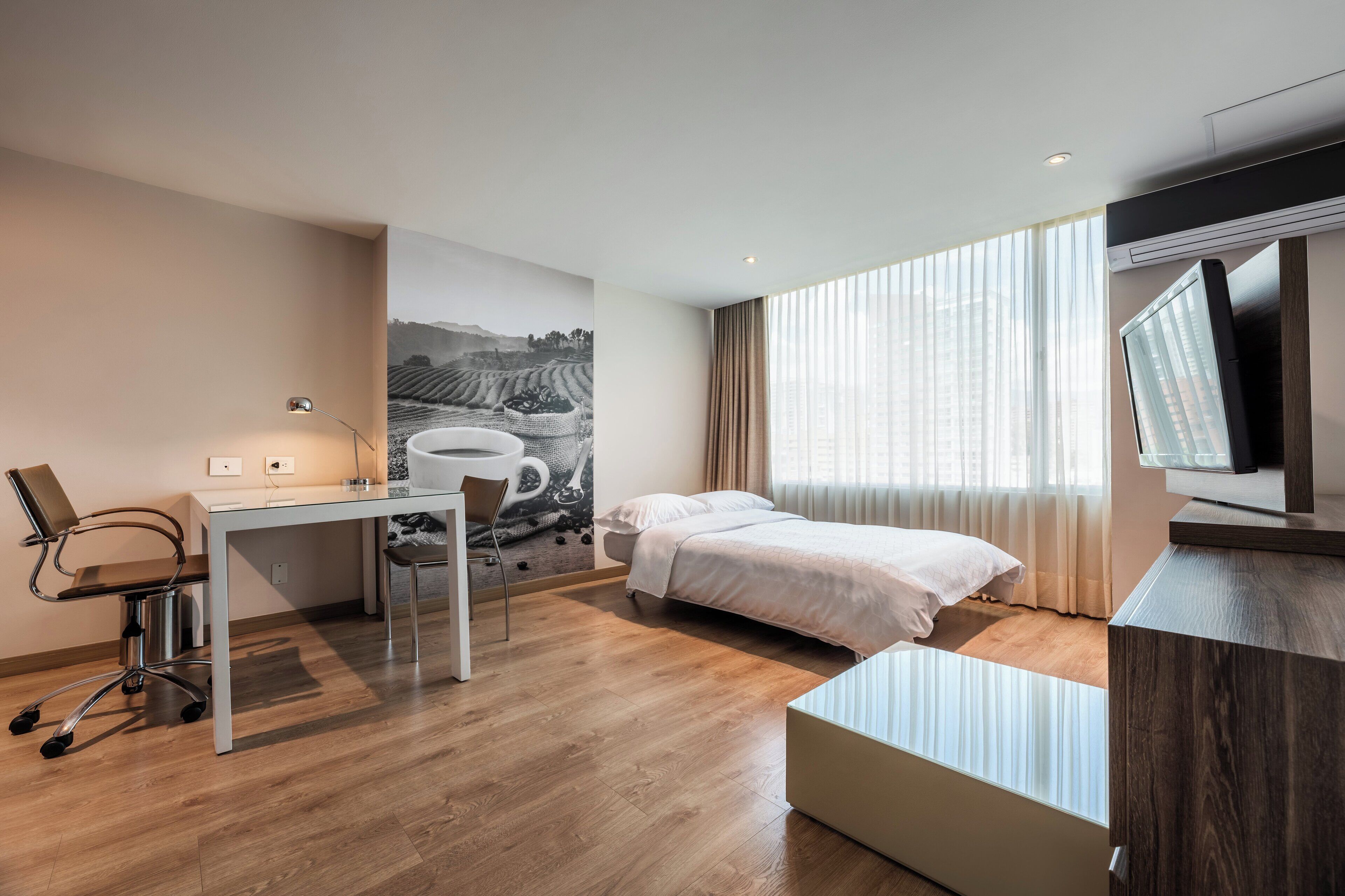 Et moderne hotelværelse med seng, skrivebord, stol og et stort vindue med gardiner.