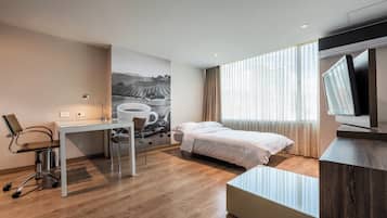 Et moderne hotelværelse med seng, skrivebord, stol og et stort vindue med gardiner.
