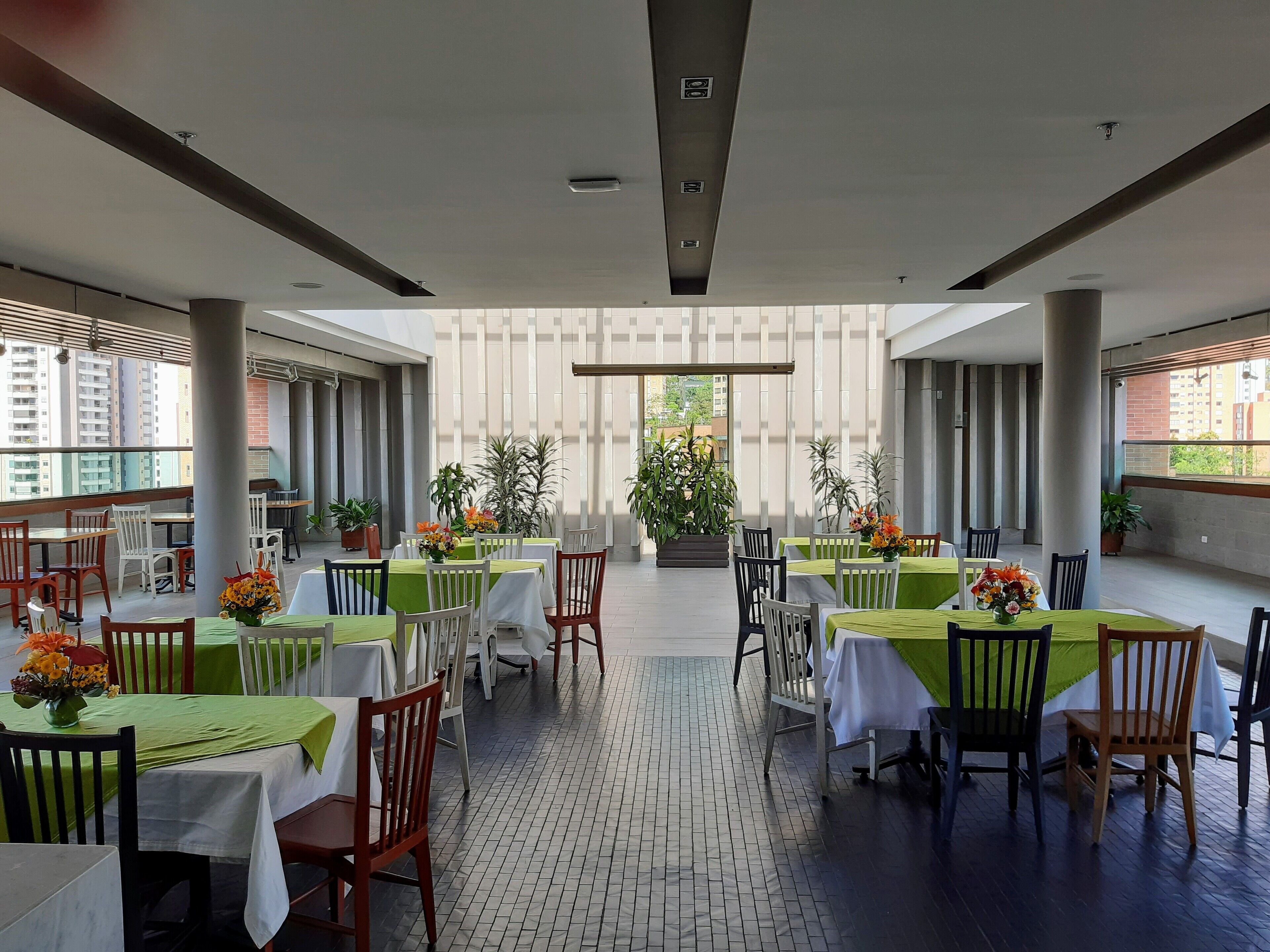 A spacious outdoor dining area with multiple tables and chairs, surrounded by potted plants and a view of buildings.