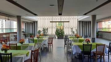 A spacious outdoor dining area with multiple tables and chairs, surrounded by potted plants and a view of buildings.