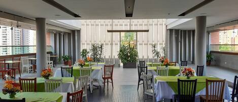 A spacious outdoor dining area with multiple tables and chairs, surrounded by potted plants and a view of buildings.