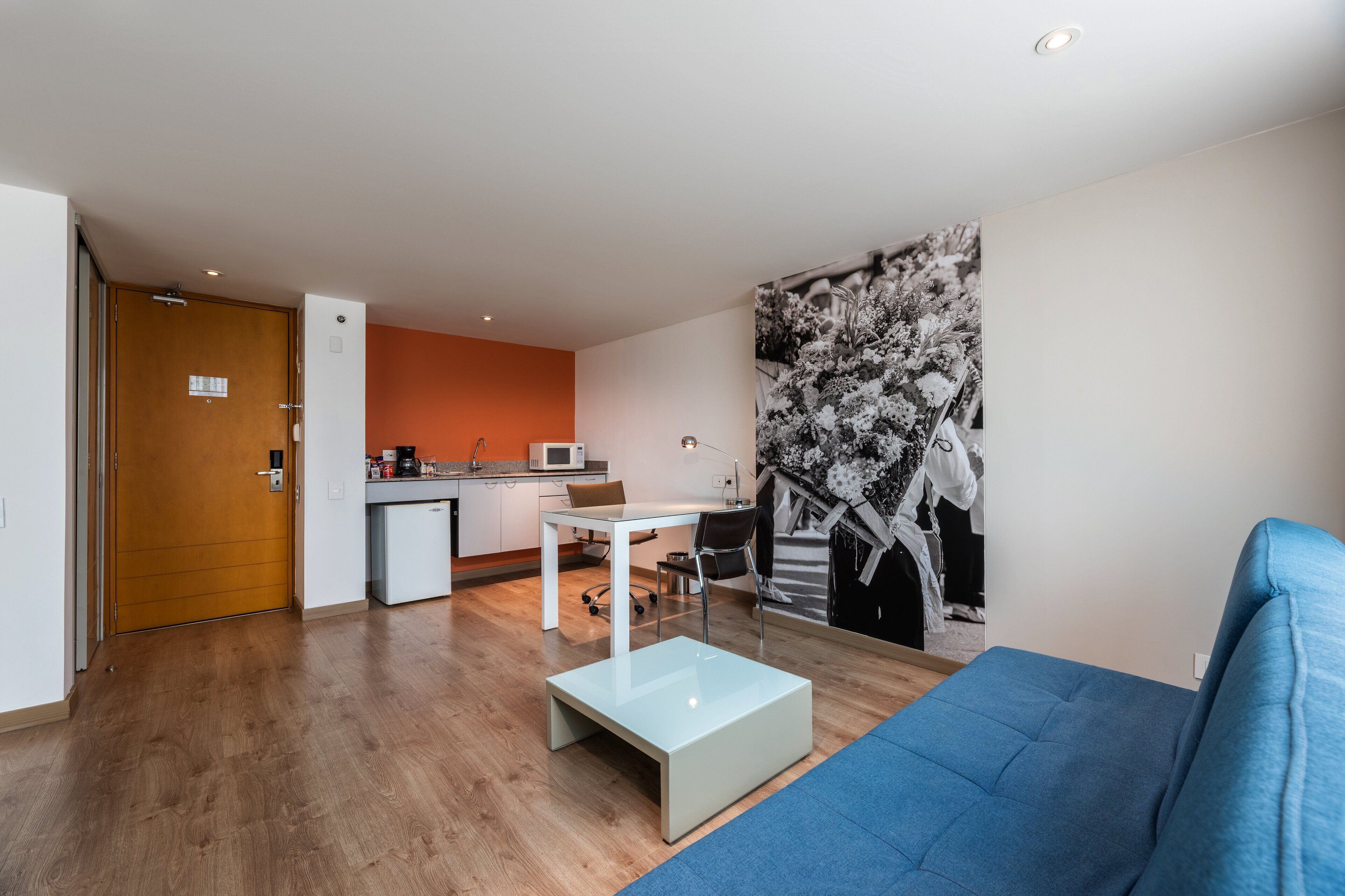 A modern living room with a blue sofa, a glass coffee table, and a kitchen area with a microwave and a small dining table.