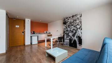 A modern living room with a blue sofa, a glass coffee table, and a kitchen area with a microwave and a small dining table.