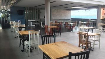 A spacious indoor dining area with multiple wooden tables and chairs, a large pillar, and a view of the ocean through large windows.