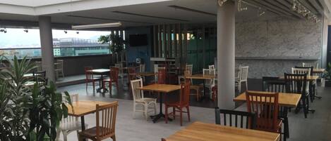 A spacious outdoor dining area with multiple wooden tables and chairs, large windows offering views of the cityscape, and a flat roof structure.