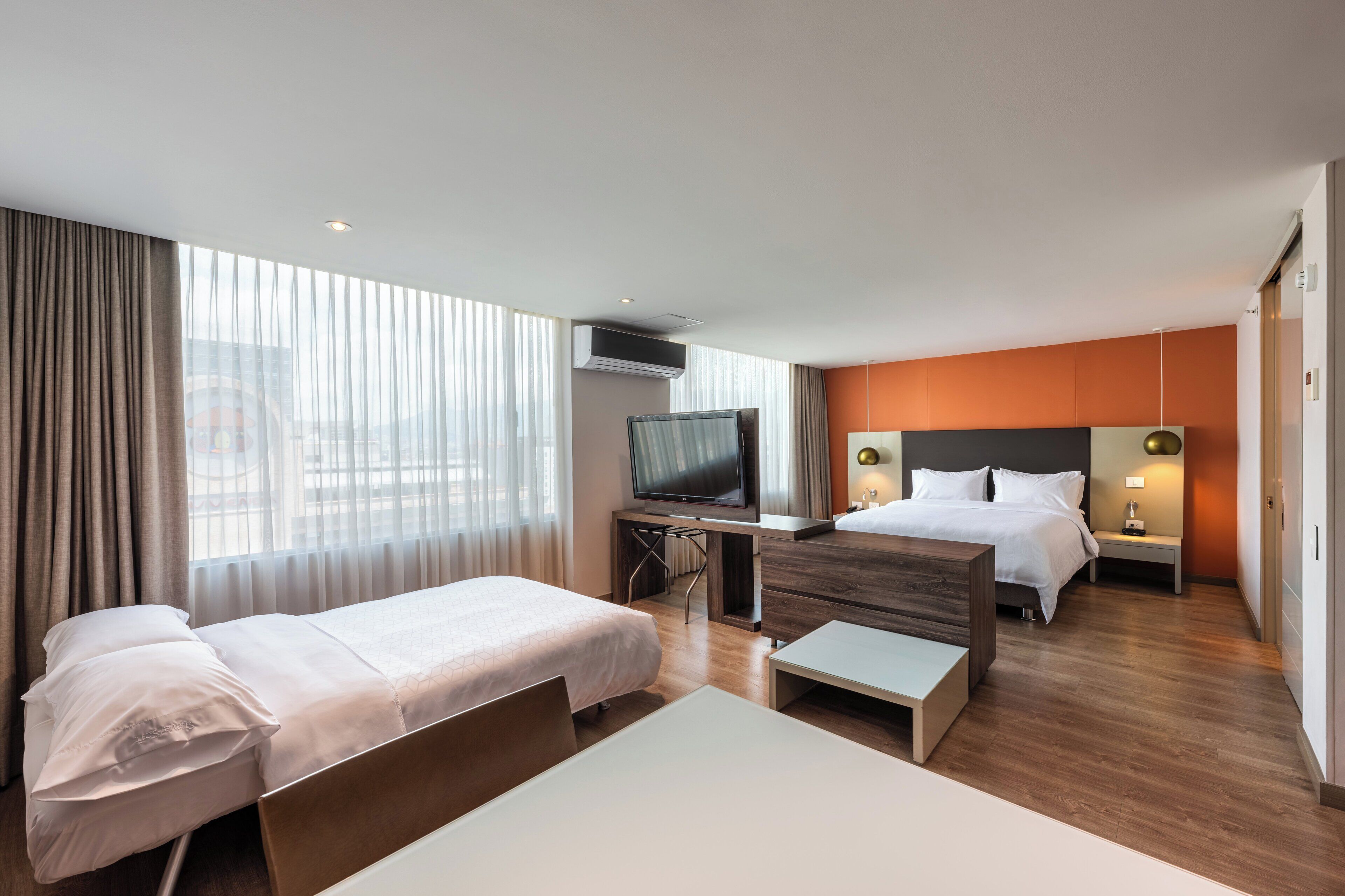 Habitación de hotel con dos camas, un escritorio con televisor y vista a la ciudad a través de amplios ventanales.