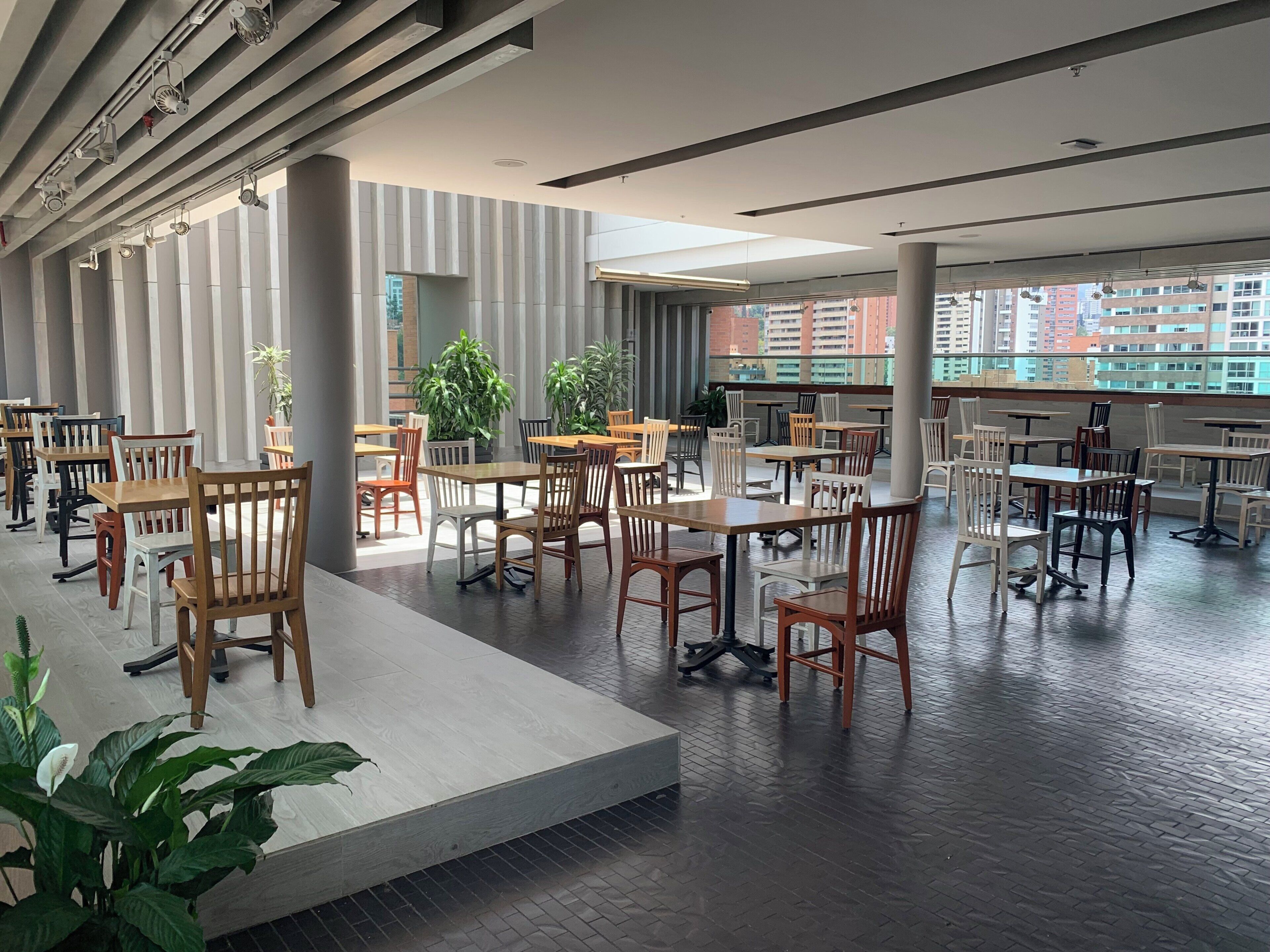 A spacious indoor dining area with multiple tables and chairs, large windows, and potted plants.