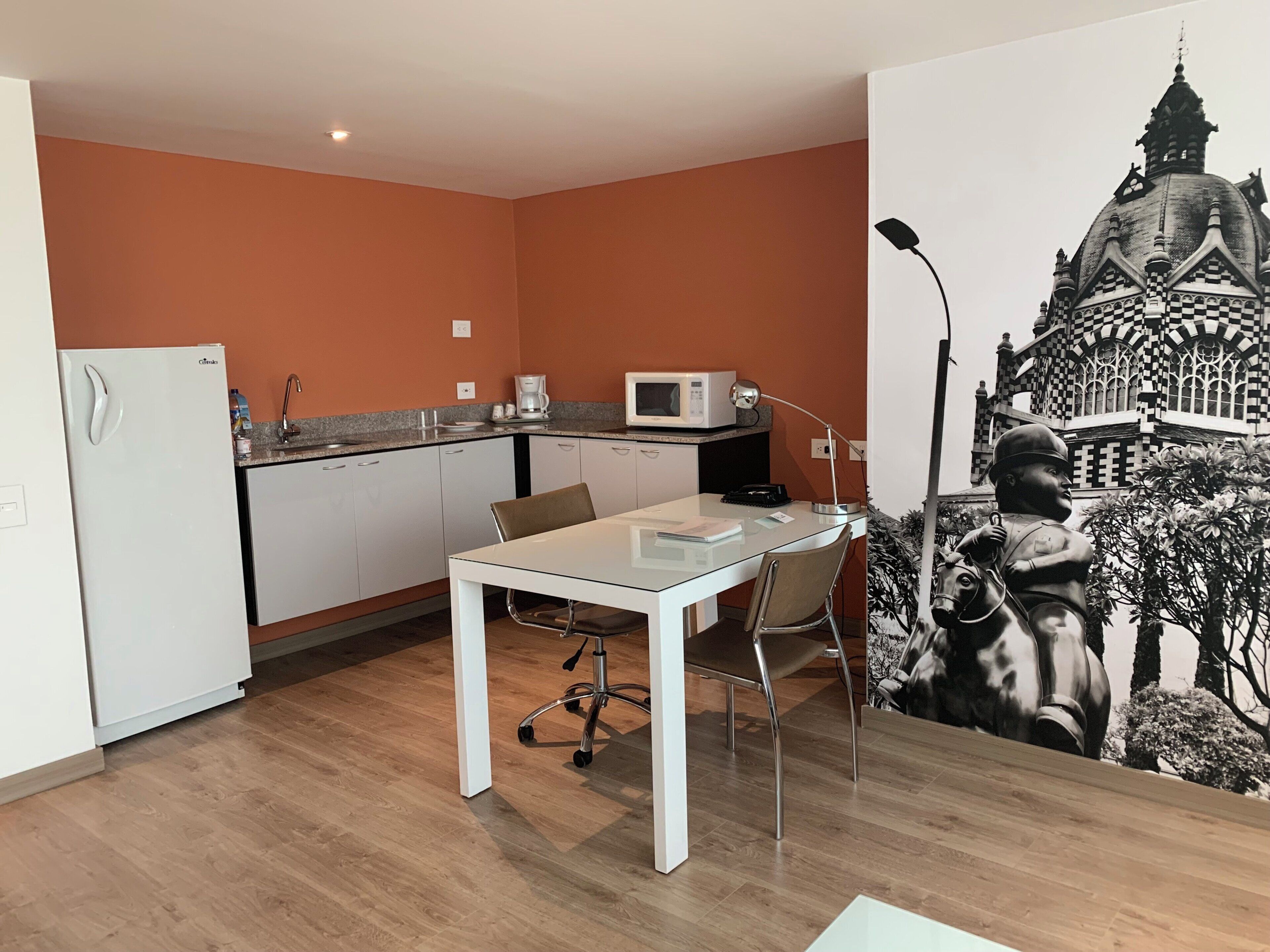 A compact kitchen with a microwave, coffee maker, and a refrigerator. There is a dining table with chairs and a large wall mural.