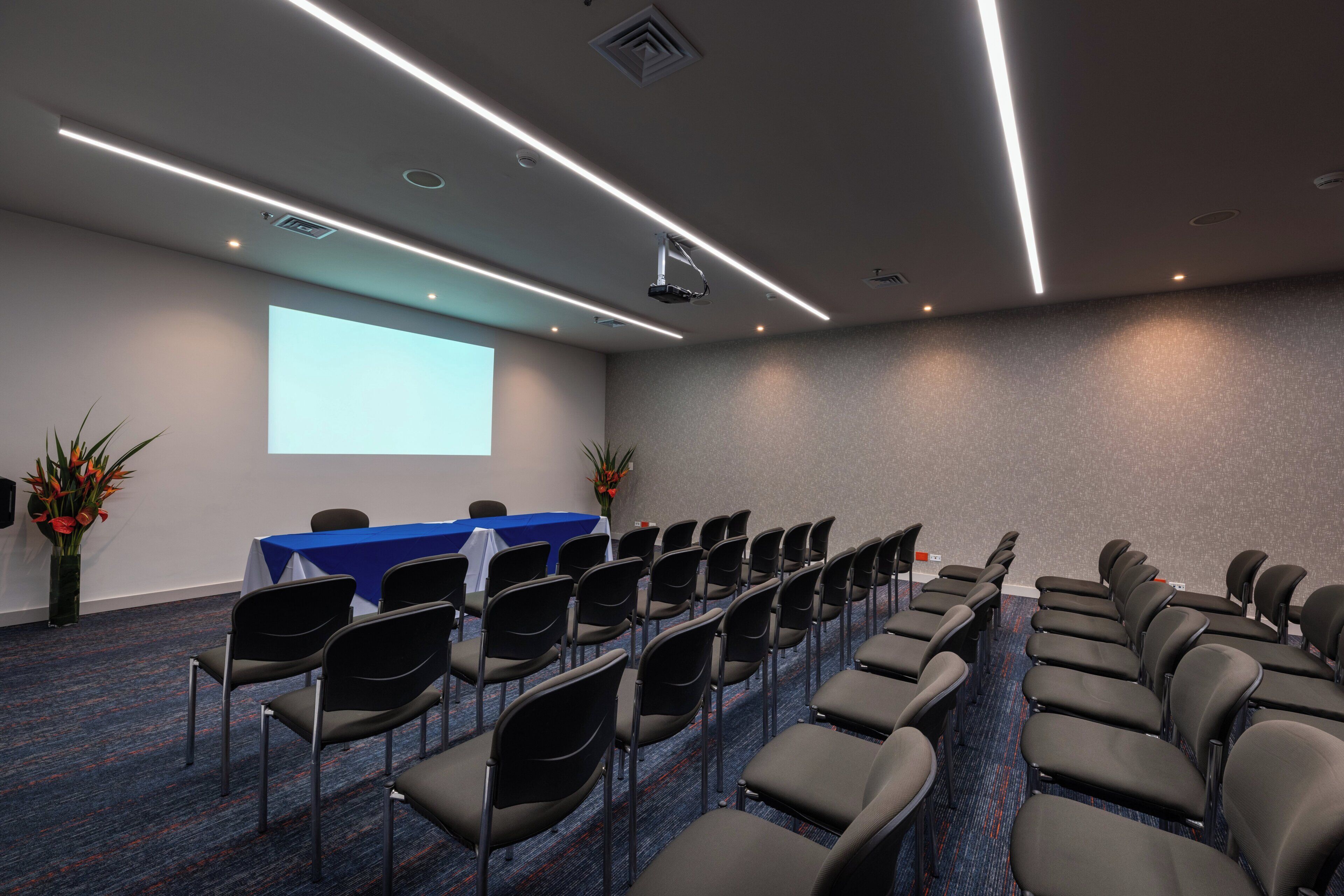 A conference room with a long table, rows of chairs, a projector screen, and a carpeted floor.