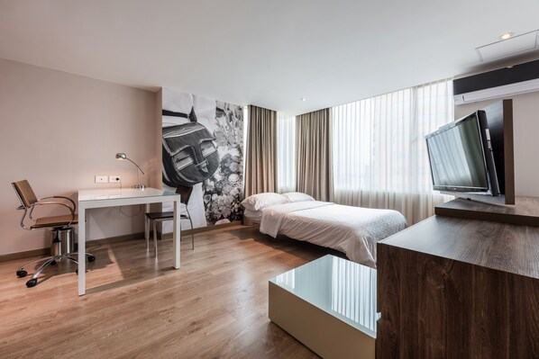 A modern hotel room with a wooden floor, a desk with a chair, a bed with white bedding, a TV on a wooden stand, and a large window with curtains. - Holiday Inn Express Medellin by IHG (Medellín)