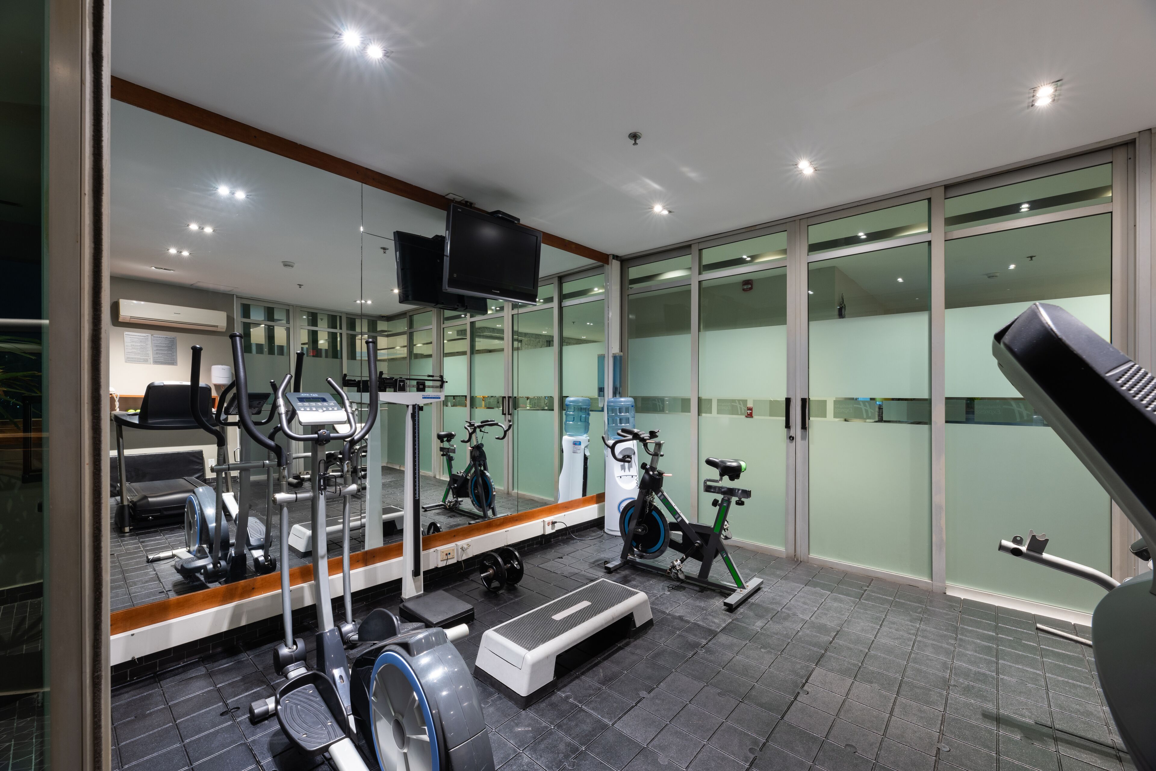 Uma academia moderna com diversos equipamentos de exercício, um espelho grande e uma televisão.