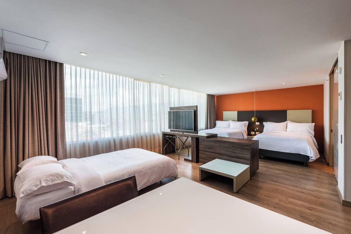 Habitación de hotel con dos camas, un escritorio, un televisor y un amplio ventanal con cortinas.