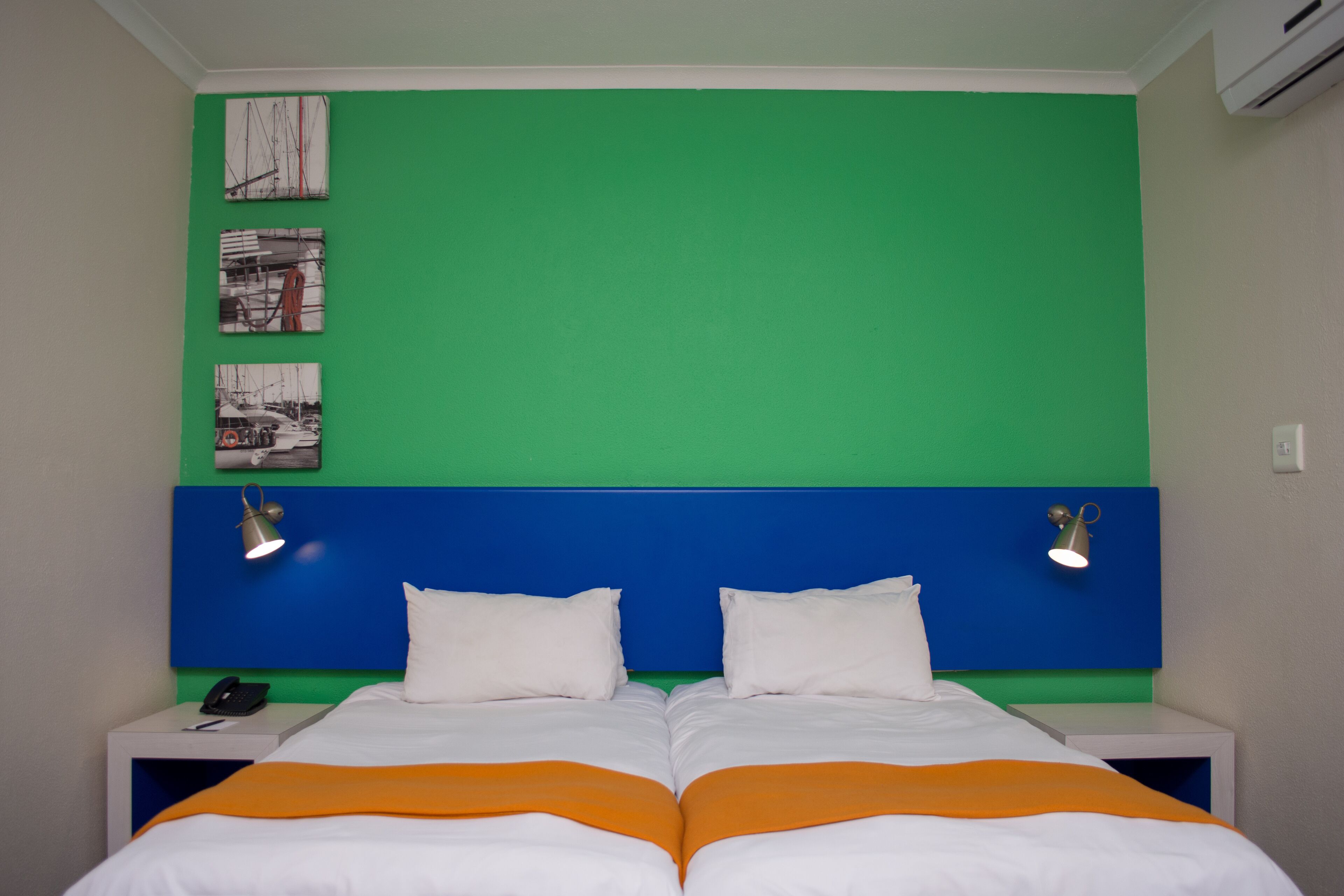 A double bed with white and orange bedding, two bedside tables with lamps, and a green wall with three framed pictures.