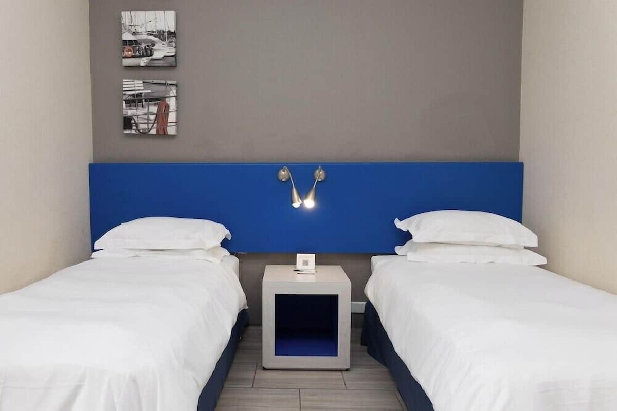 A hotel room with two beds, a blue headboard, a small bedside table with a lamp, and two framed pictures on the wall.