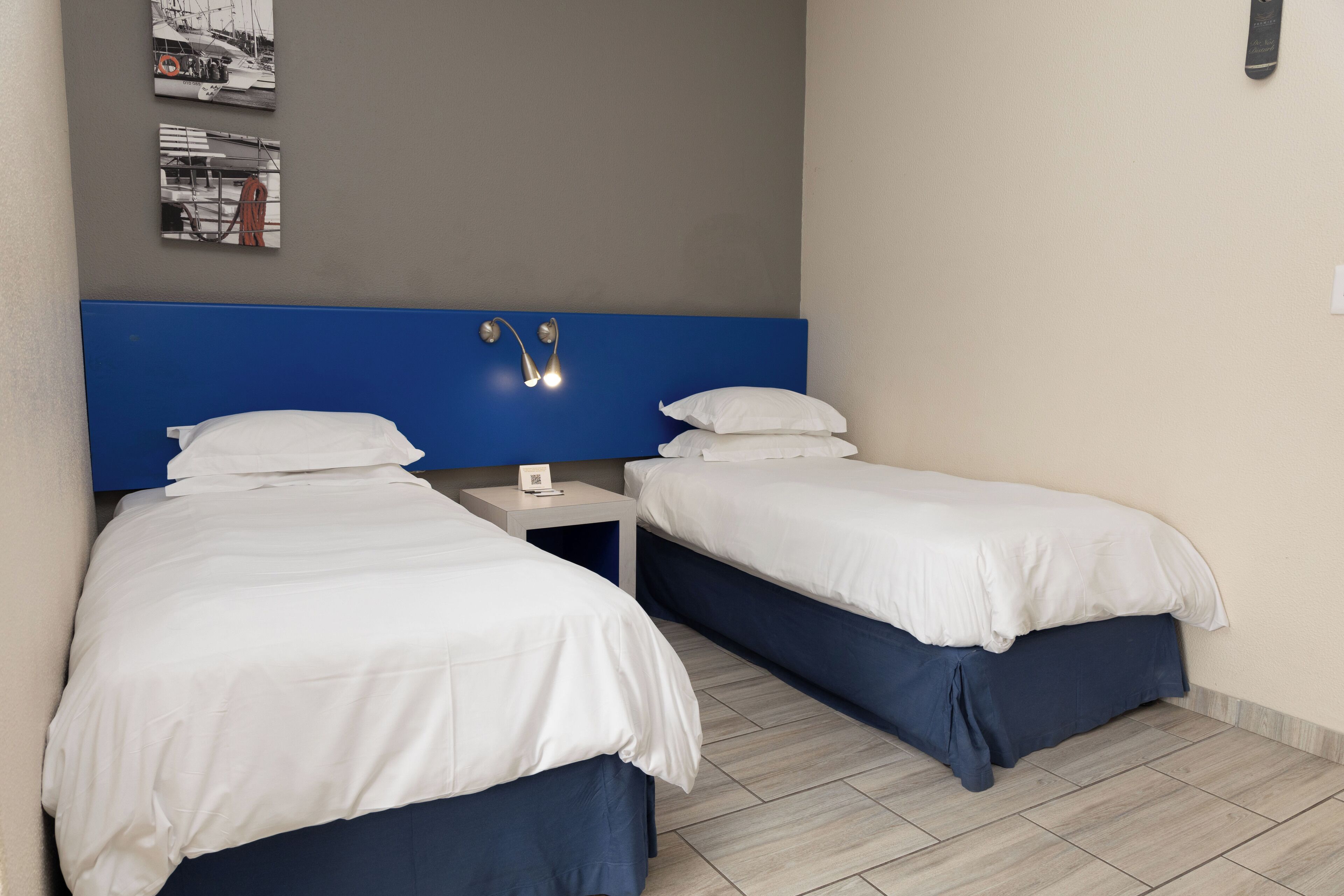 A hotel room with two beds, a blue headboard, a small bedside table with a lamp, and a wall-mounted shelf with books.