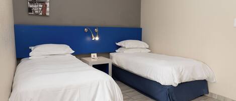 A hotel room with two beds, a blue headboard, a small bedside table with a lamp, and a wall-mounted shelf with books.