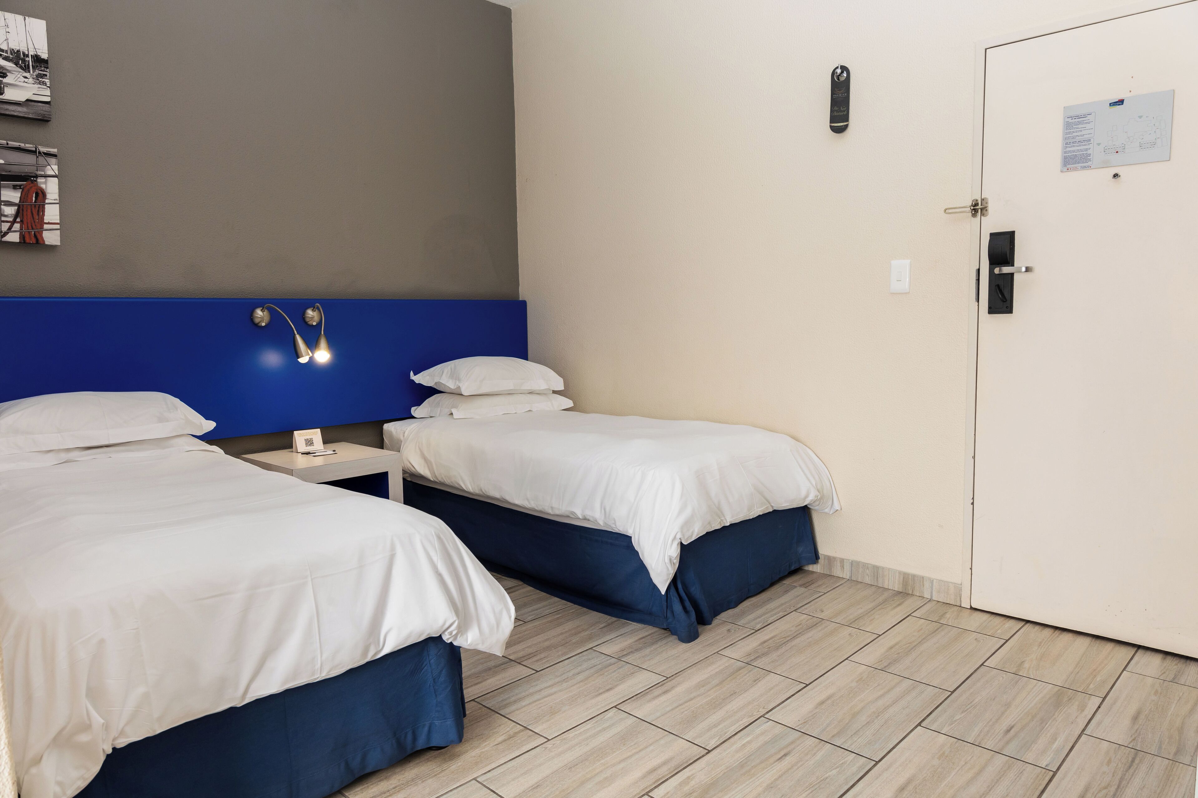 A hotel room with two beds, a nightstand, a wall-mounted light, a door, and a bulletin board.