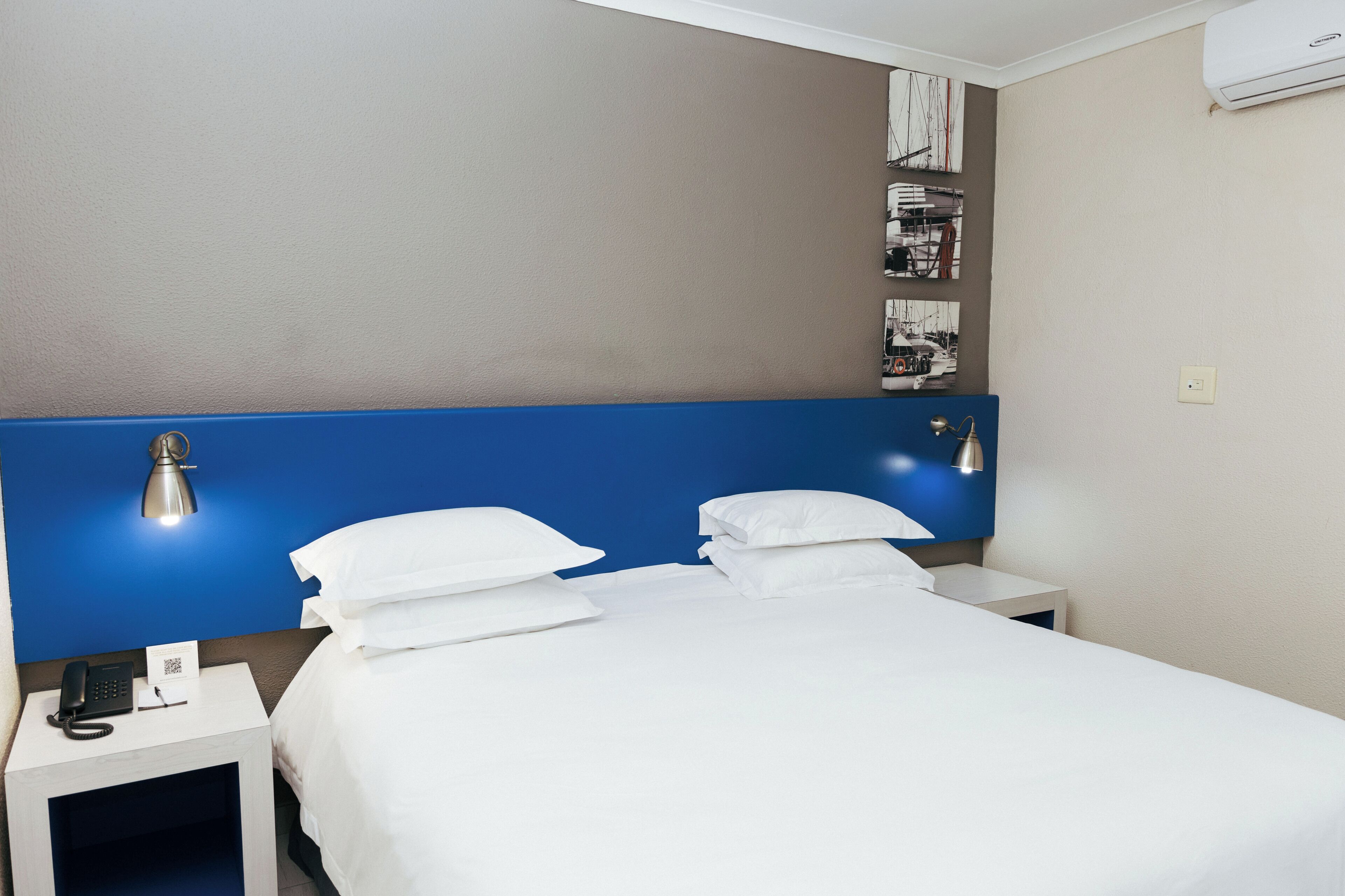 A hotel room with a bed, white pillows, a bedside lamp, a phone, and a small table with a lamp.