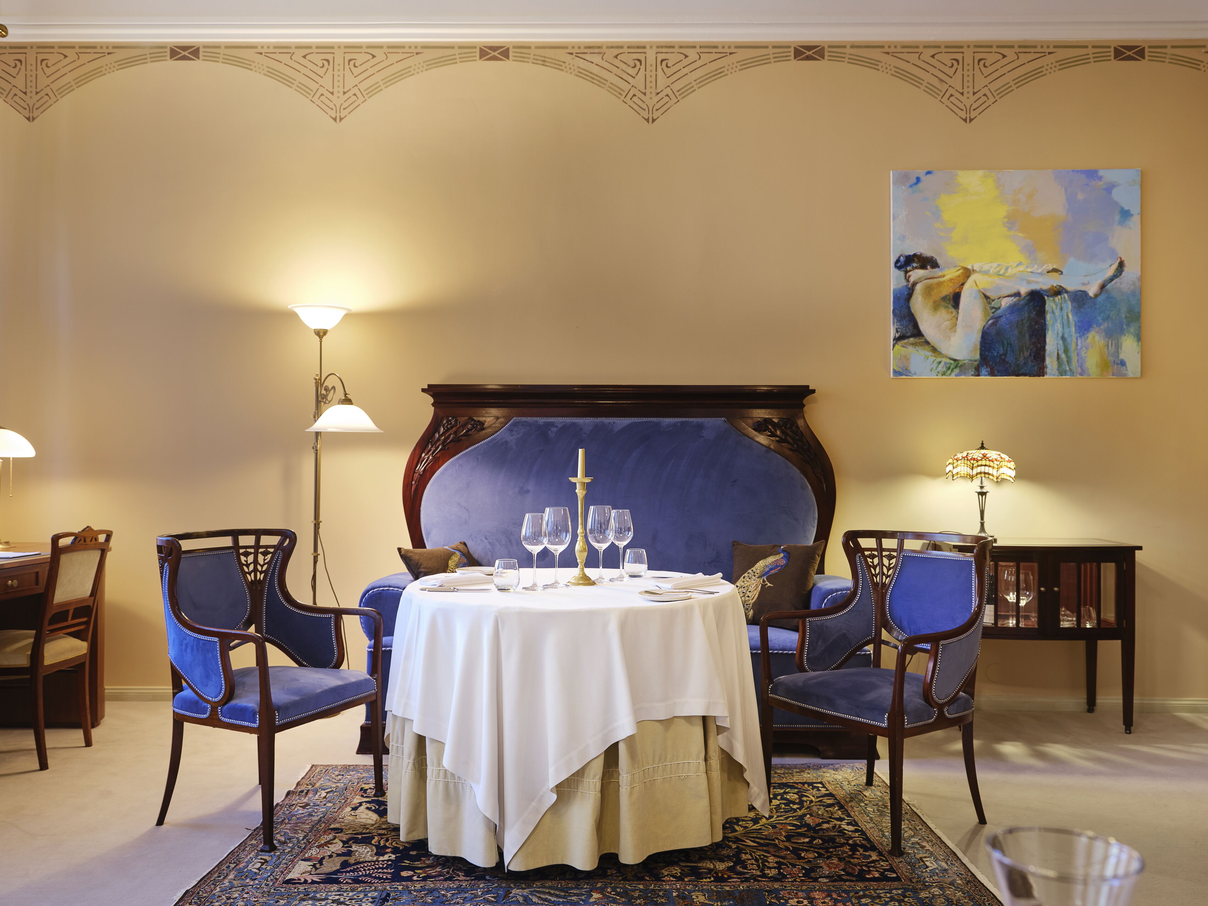 A dining area with a round table set for four, two blue upholstered chairs, a decorative headboard, and a large painting on the wall.