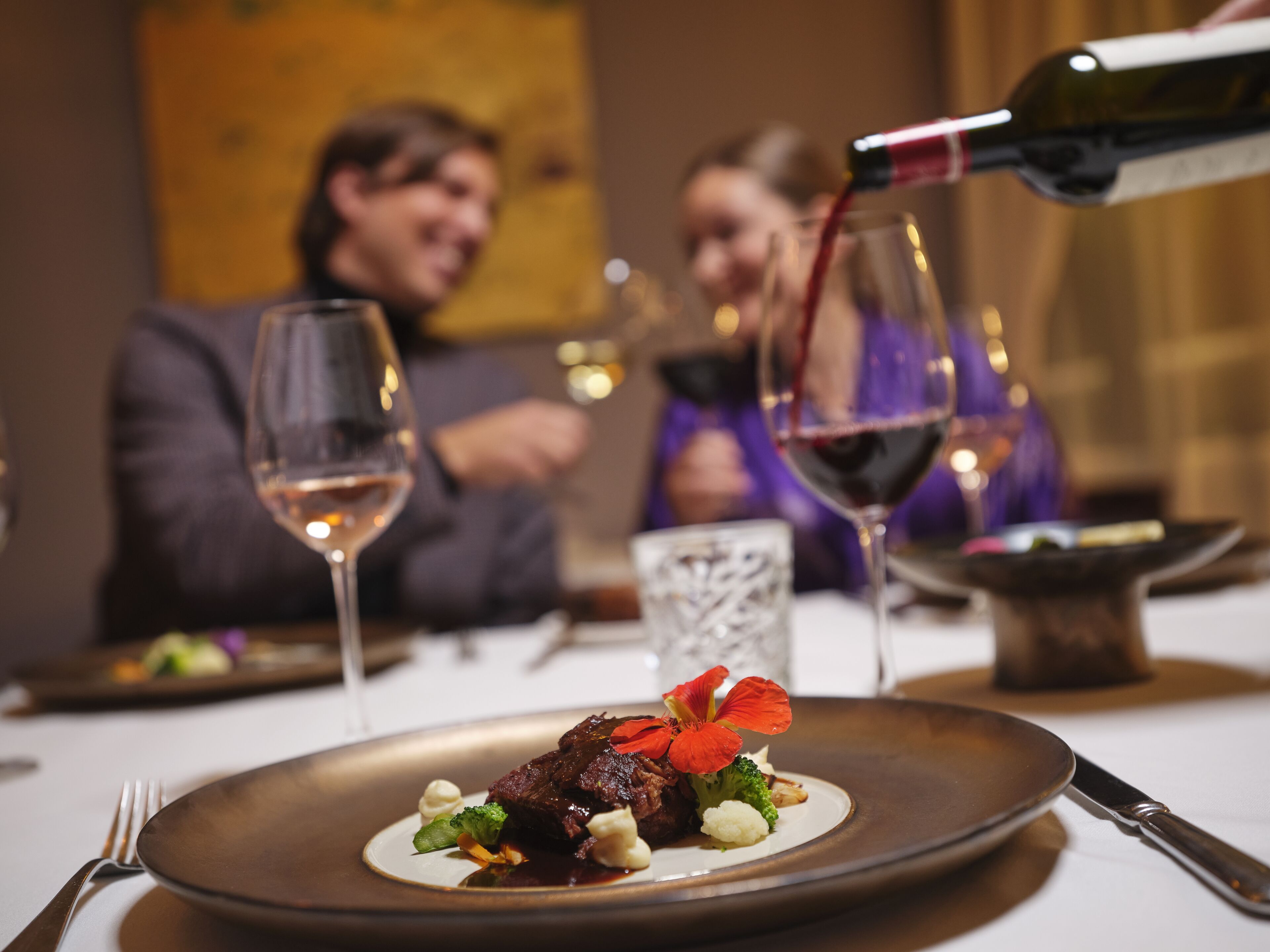 Mesa de jantar com prato de comida, copos de vinho e uma garrafa de vinho sendo servida.