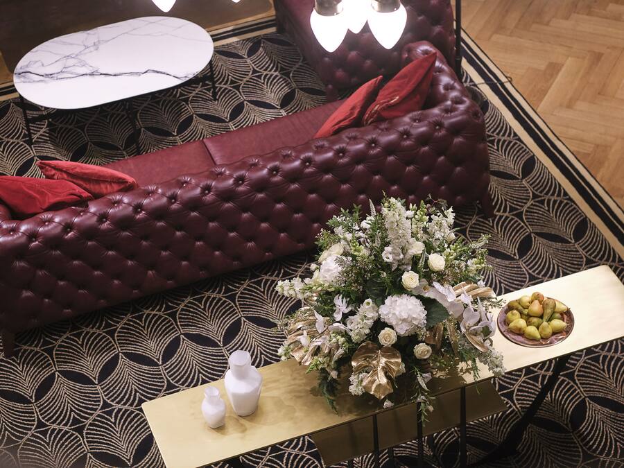 A plush red sofa with red cushions, a small white vase, and a floral arrangement on a table with a patterned floor.