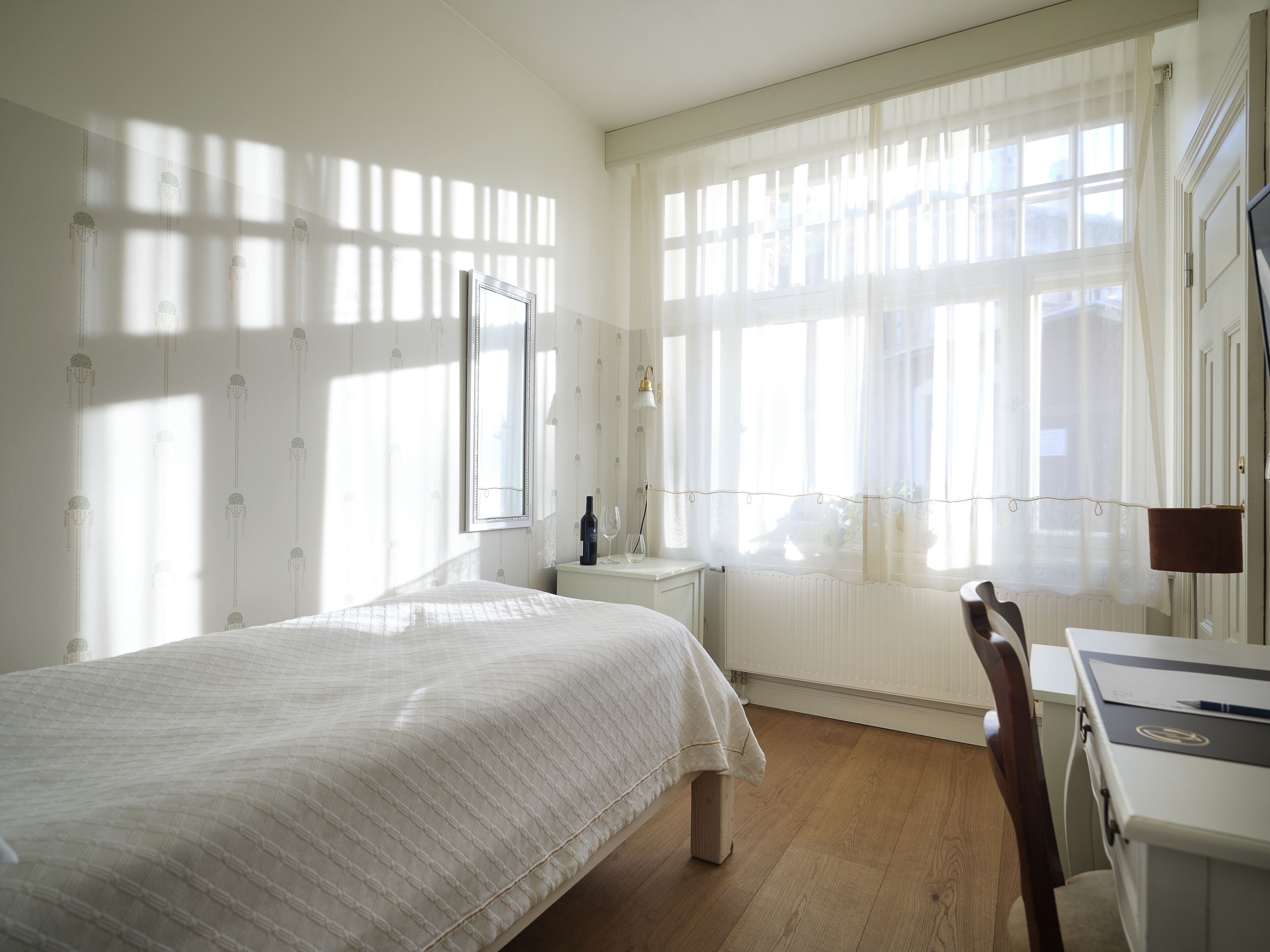 A bedroom with a bed, a desk, and a chair. There is a window with curtains and a radiator.