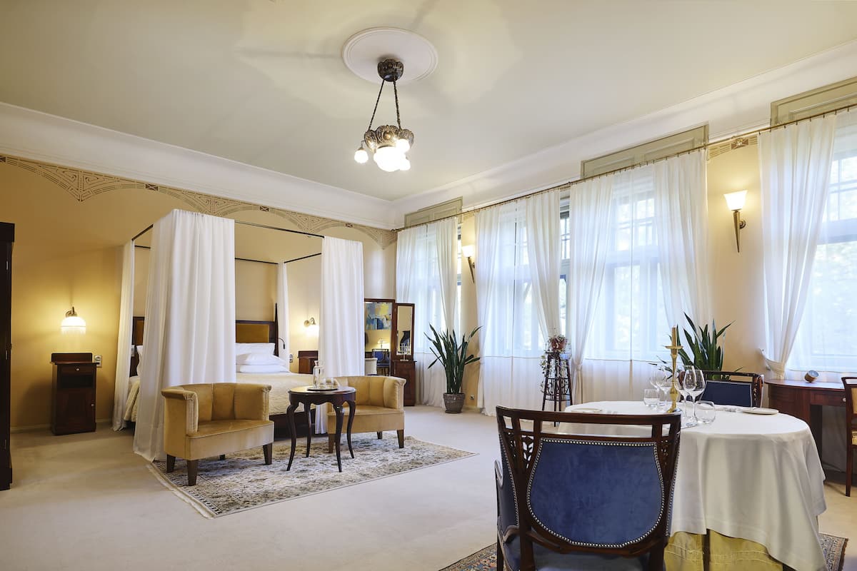 Une chambre d’hôtel dotée d’un lit à baldaquin, d’un coin salon avec un canapé et des fauteuils, d’une table à manger avec des chaises et d’une grande fenêtre avec des rideaux.