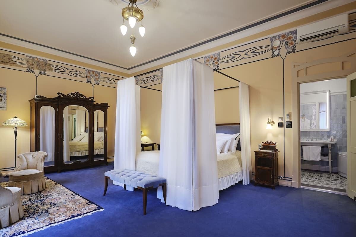 Une chambre à coucher avec un lit à baldaquin, un coin salon avec un banc, un grand miroir et une salle de bain avec un lavabo et un miroir.
